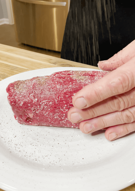 Beef being seasoned with salt and pepper.