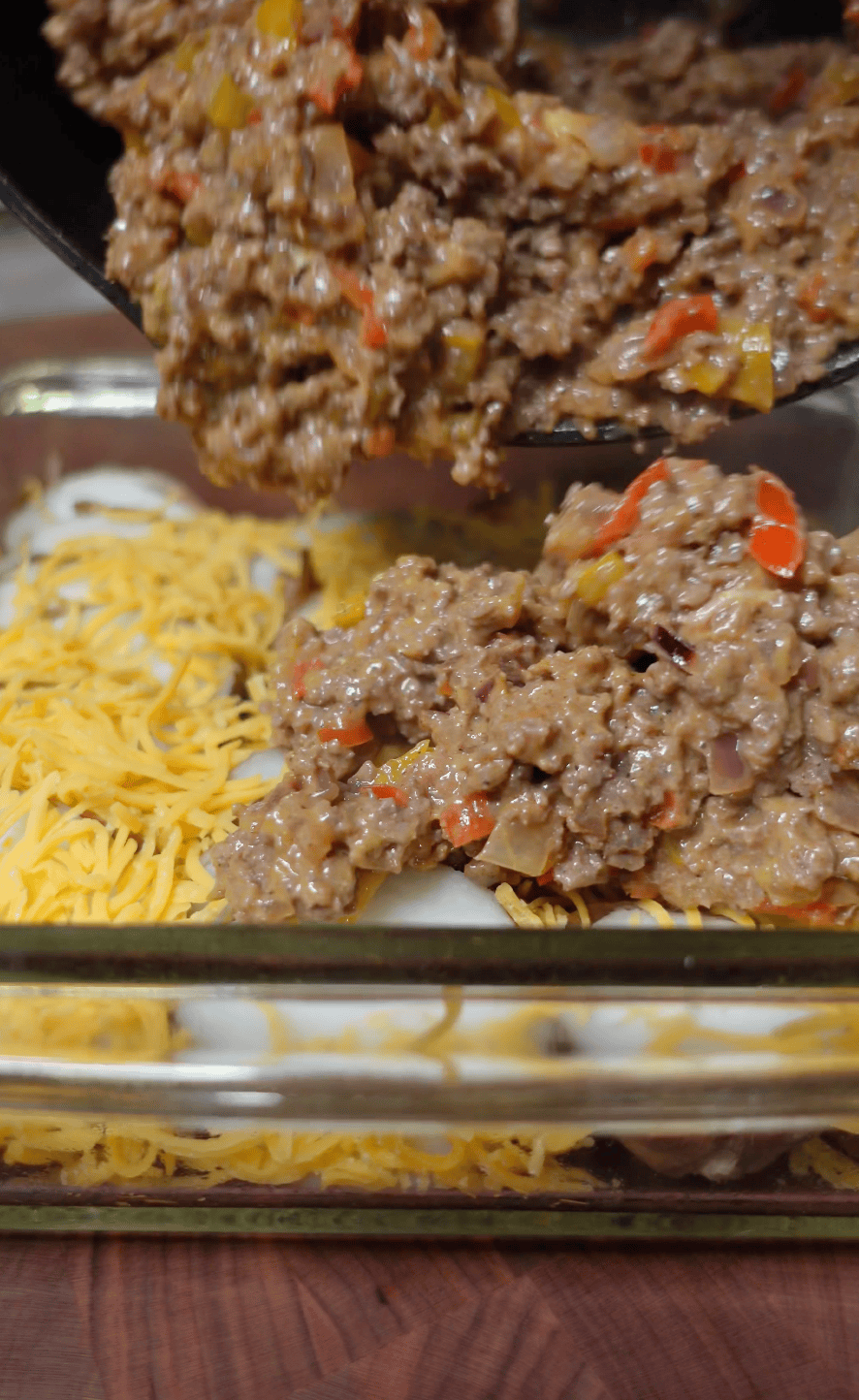A glass baking dish with potatoes and shredded cheddar in the bottom. Ground beef is being added over the top. 