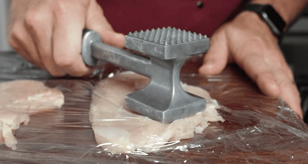 A hand holding a meat mallet and pounding chicken breast with plastic wrap on top. 