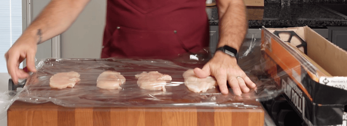 A wooden cutting board with plastic wrap and chicken breasts on top. A hand putting plastic wrap on top of the chicken breasts. 