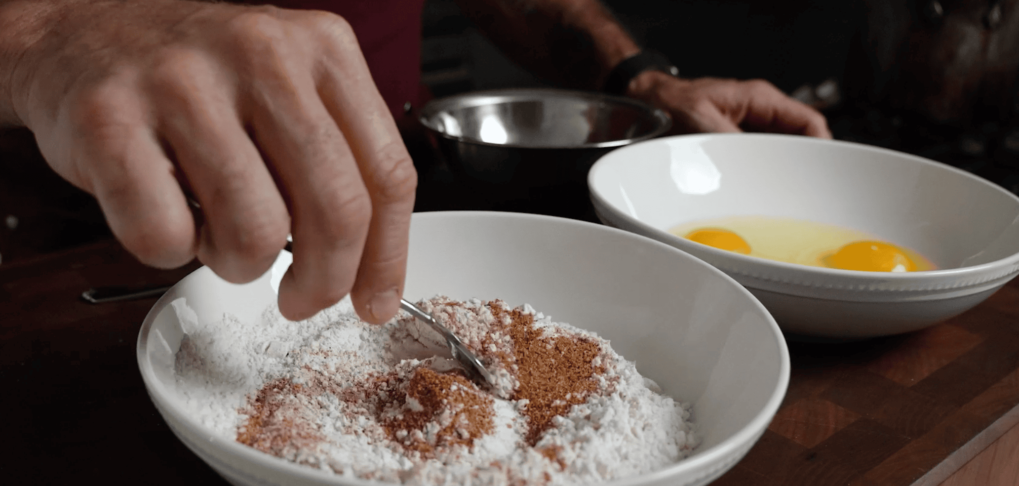 Two white bowls, one with flour mixes with a hand mixing, and the other with eggs. 