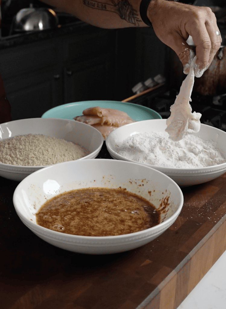 A hand dredging a chicken breast in flour, then egg, then panko breadcrumbs. 