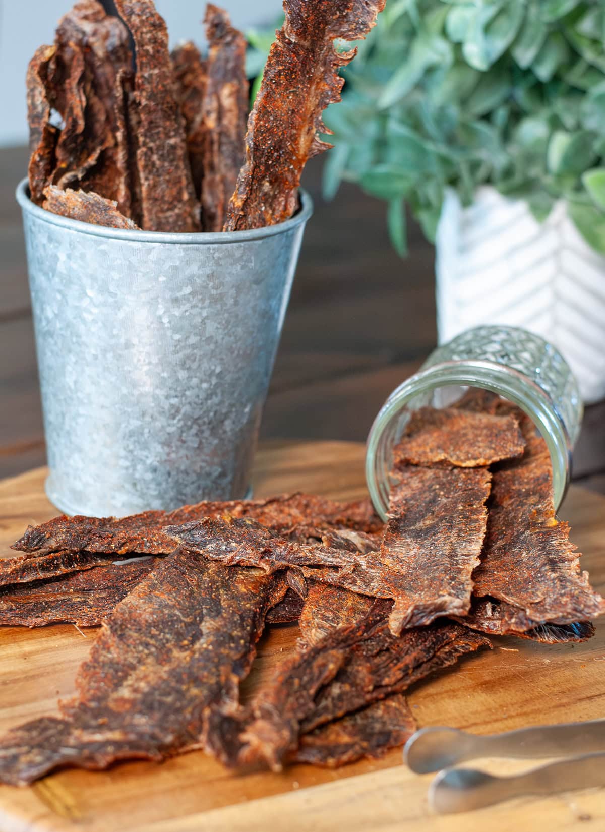 Homemade beef jerky on a wooden cutting board with tongs.