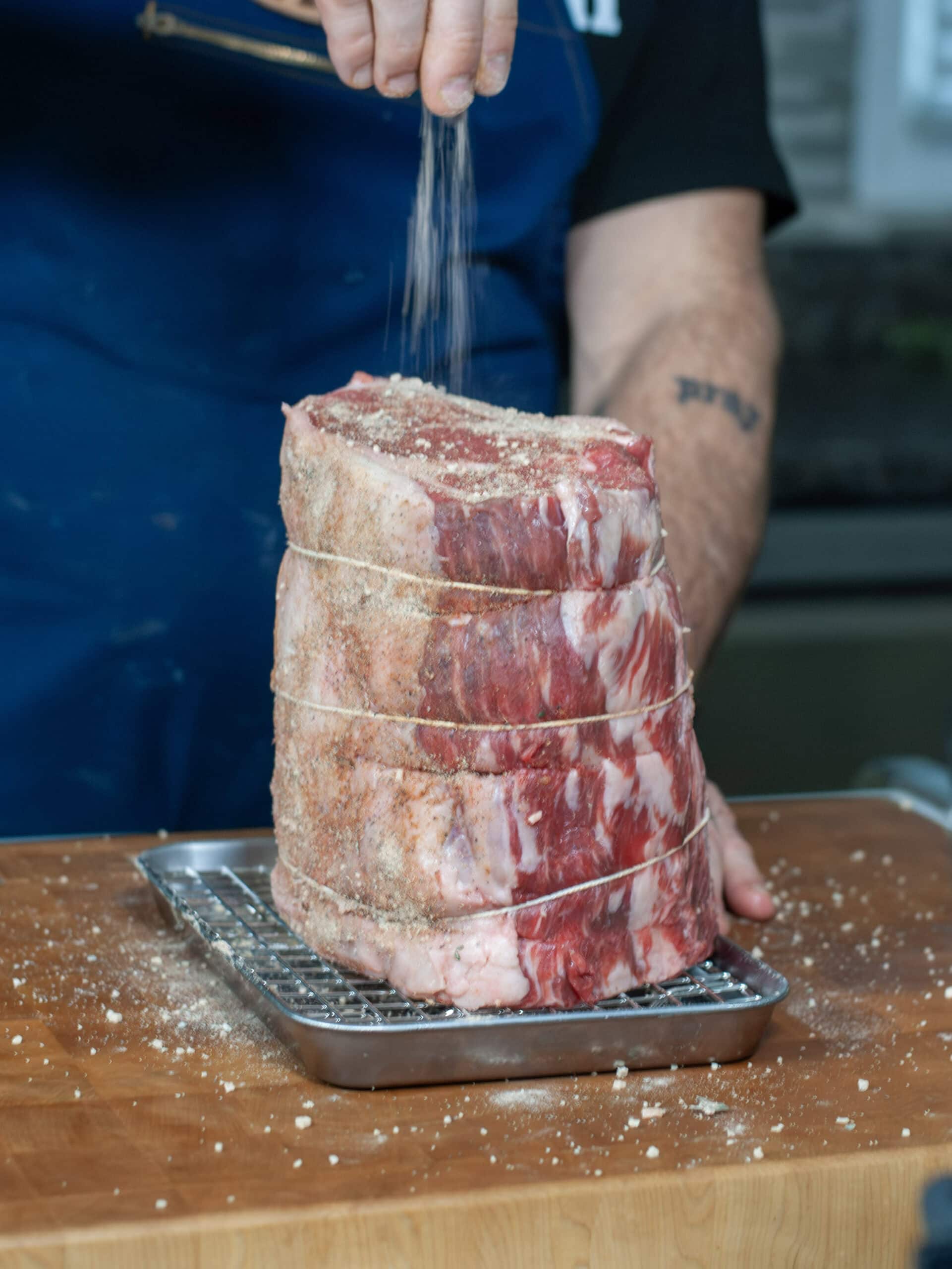 A rib roast tied with twine being seasoning with prime rib seasoning.