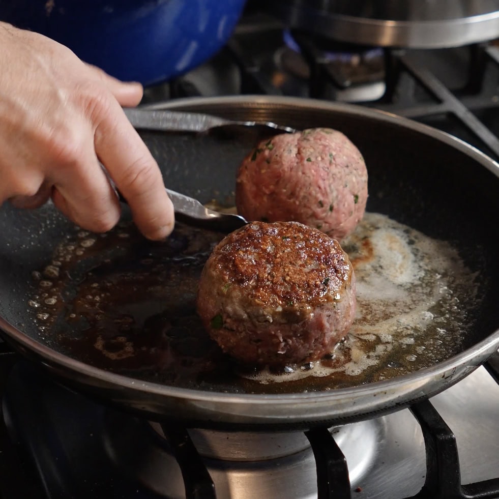 Large meatballs being cooked in a skillet.