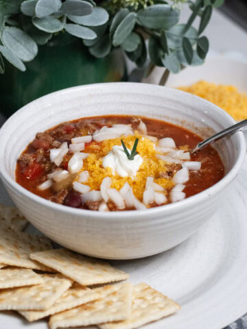 A bowl of chili with saltines, cheese and onions.
