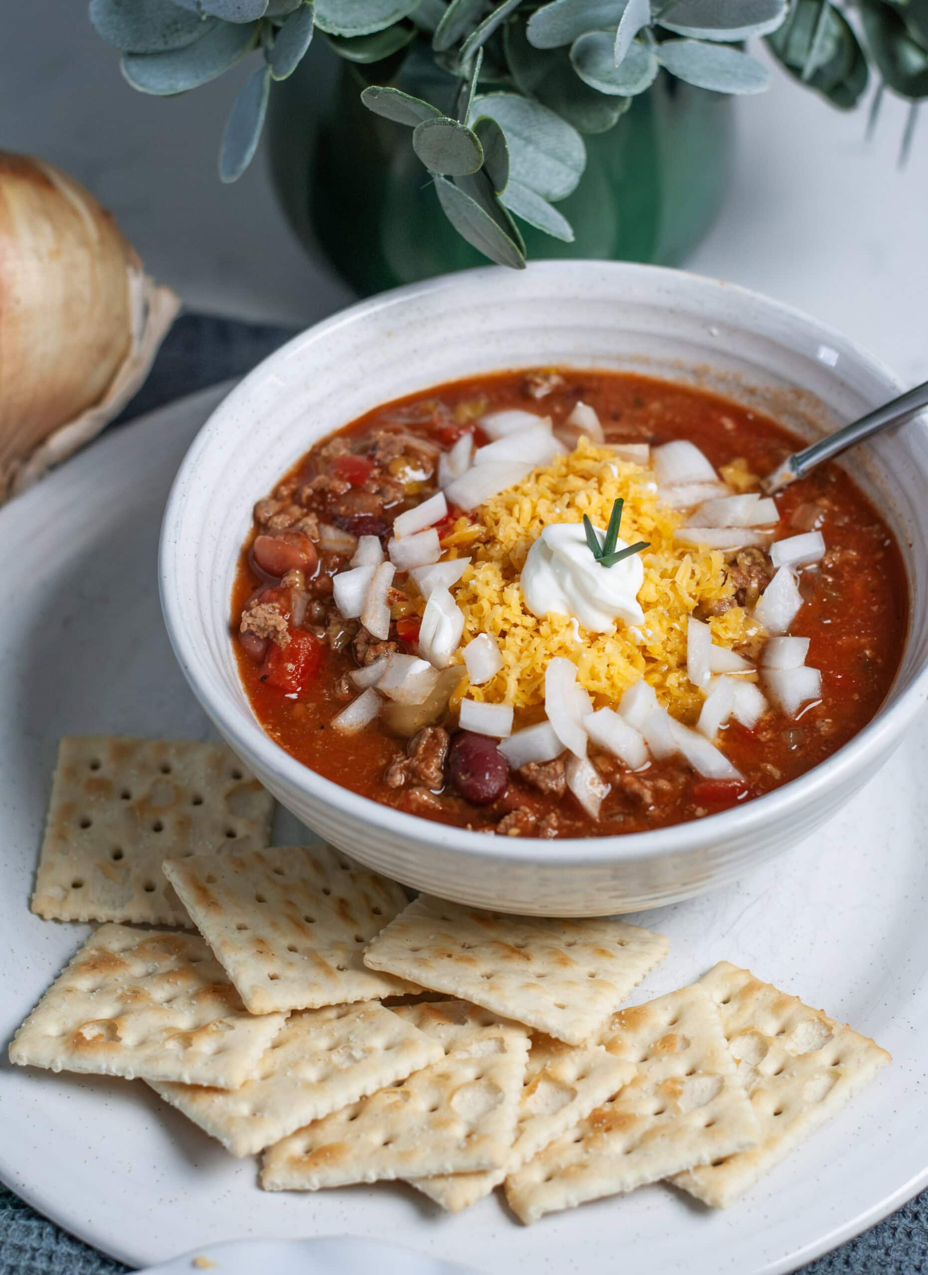 A bowl of chili with saltines, cheese and onions.