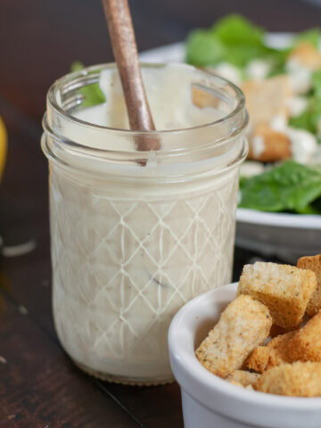 Caesar salad dressing in a glass jar next to a salad and croutons.