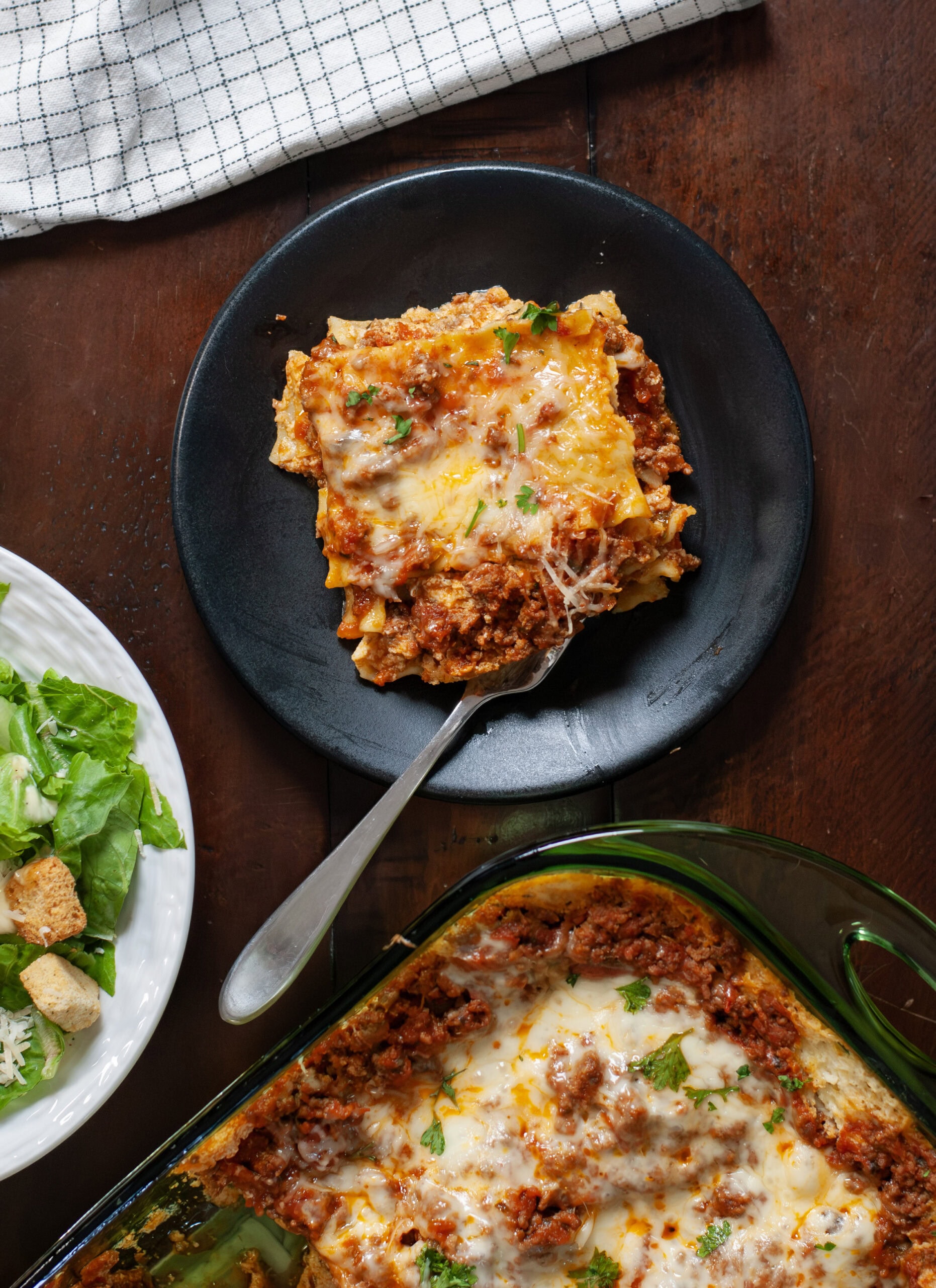 Lasagna on a plate next to caesar salad and a baking dish of lasagna.