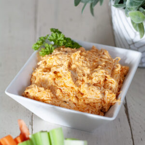 A white bowl of buffalo chicken dip next to celery and carrots.
