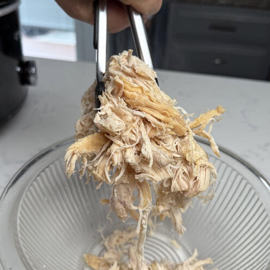 Tongs holding shredded chicken over a glass bowl.