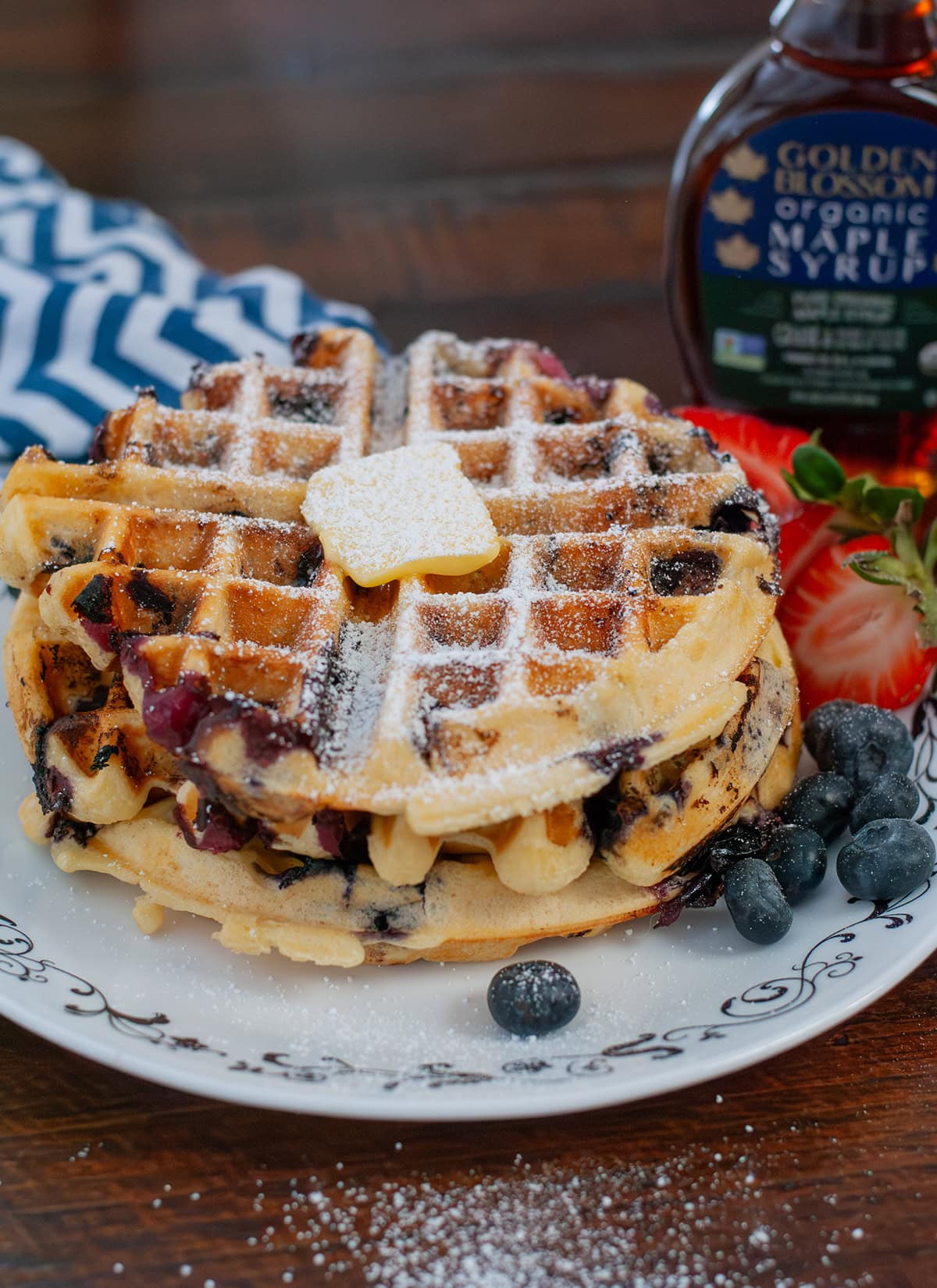 A blueberry waffle on a plate with fruit and powdered sugar.