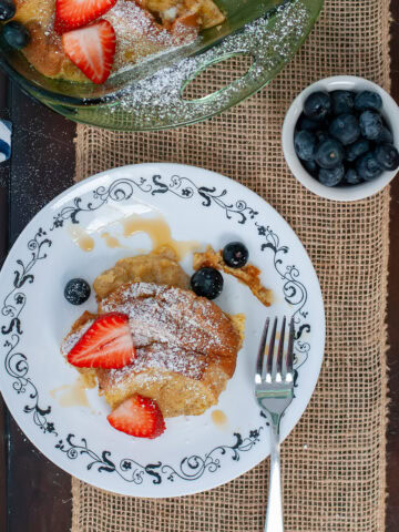 A brioche casserole on a plate with powdered sugar and strawberries and blueberries.