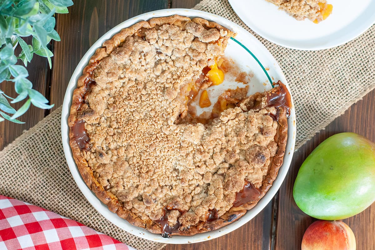 A peach mango pie with a slice on the side next to mangos and peaches.