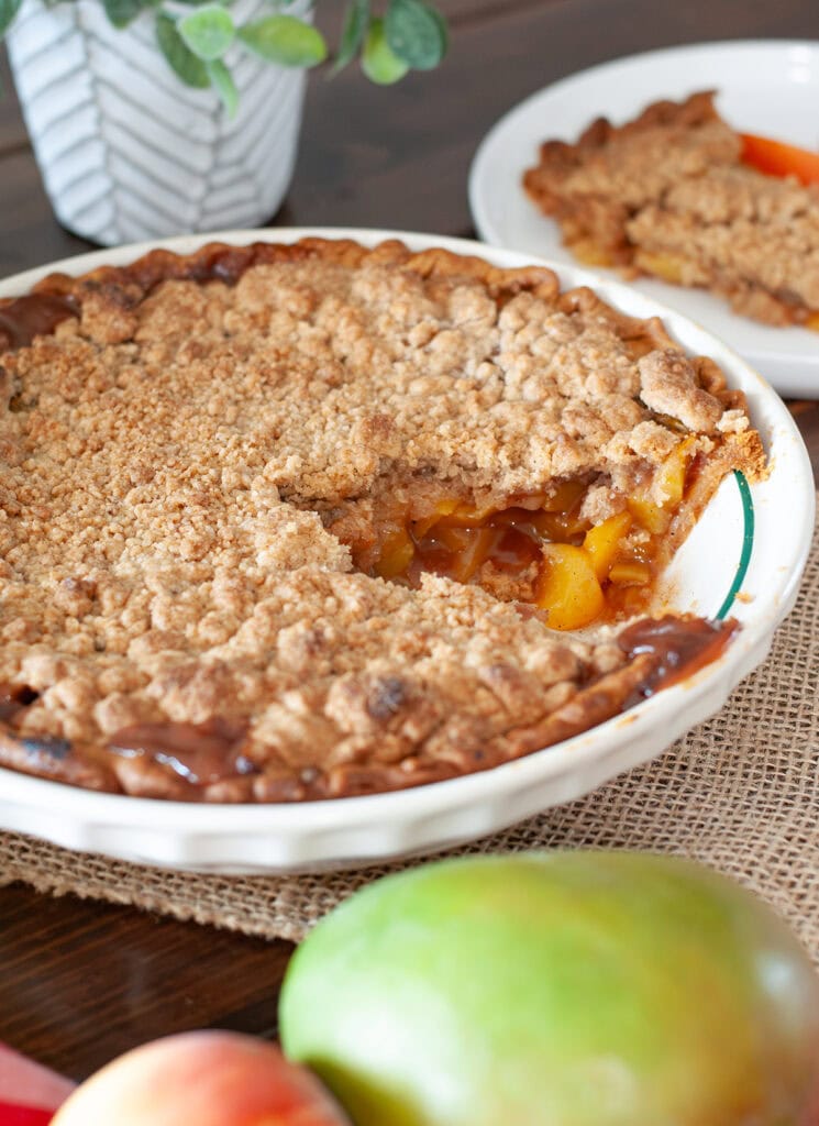 A pie with a slice missing sitting next to a mango and peaches.