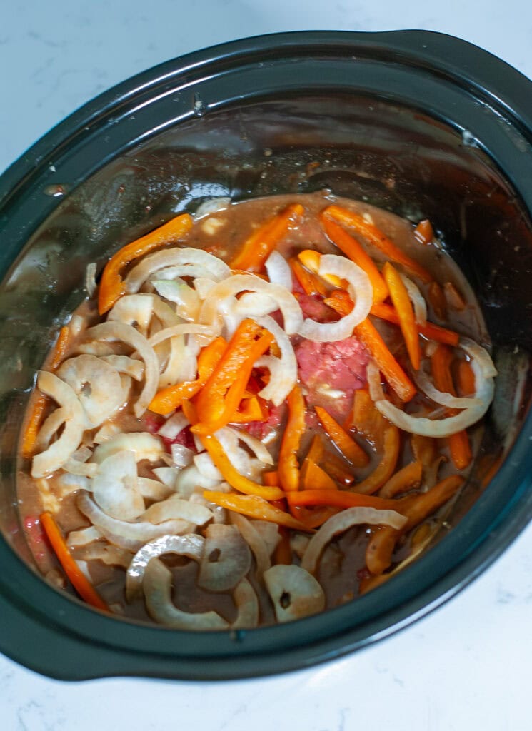 Sliced onions, peppers and cube steak in a crock pot with sauce.