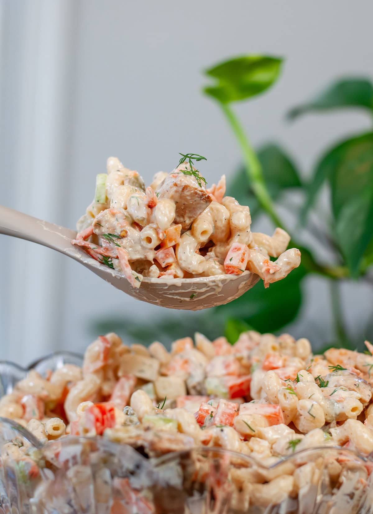 A glass bowl with a spoon of a creamy pasta salad showing diced chicken, bell peppers, and fresh dill. 