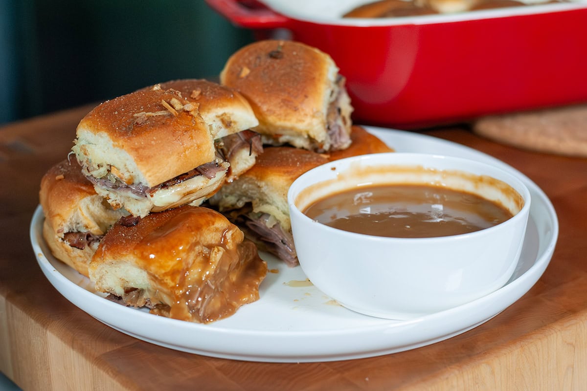 A stack of sliders on a plate beside a bowl of brown gravy. 