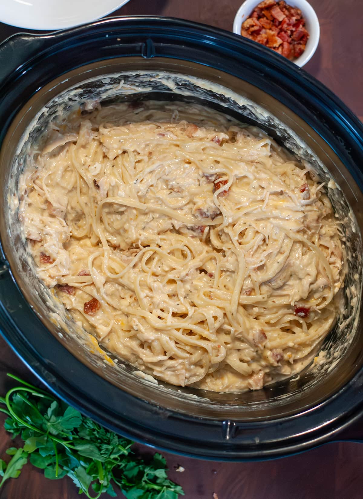 Finished chicken pasta in a crock pot next to bacon bits.