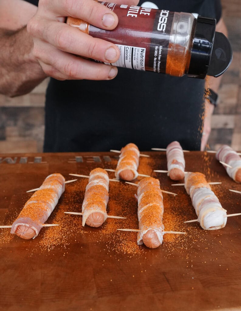 BBQ seasoning being put on bacon wrapped hot dogs before grilling.