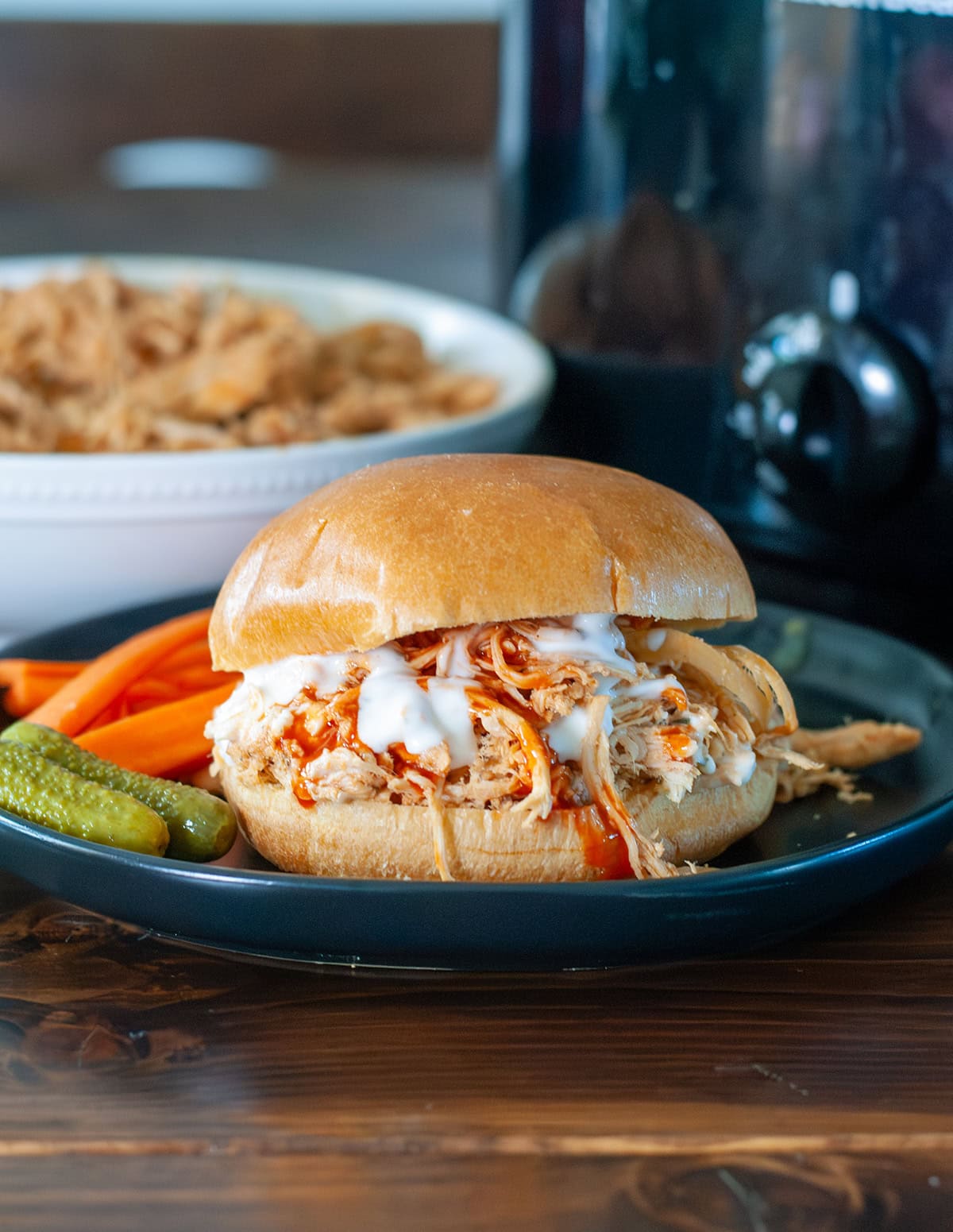 A shredded chicken sandwich with ranch dressing, hot sauce on a bun served with pickles and carrots. 