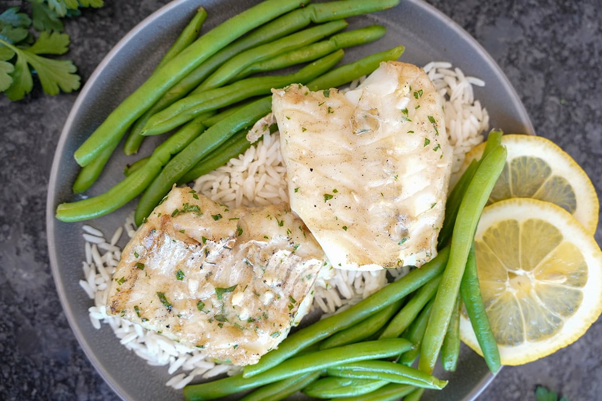 A plate of grilled cod on rice with green beans and lemon slices. 