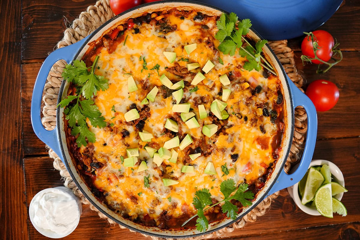 A top view of a cooked skillet Mexican beef and rice showing melted cheese, diced avocado, cilantro and small bowls of lime and sour cream. 