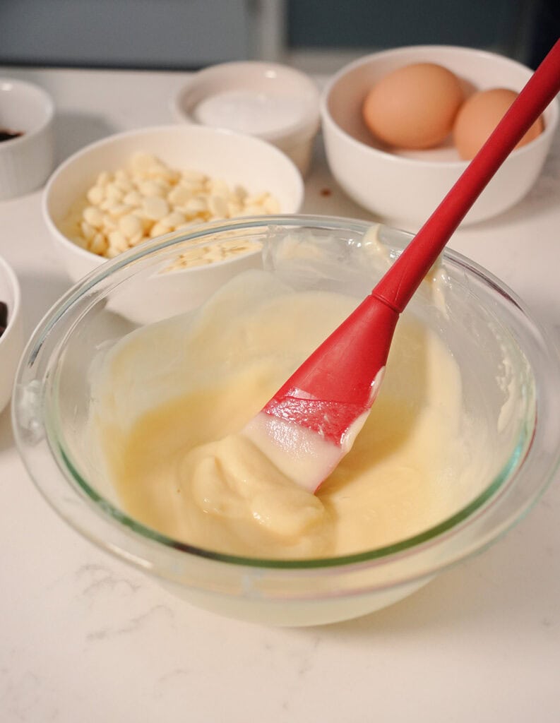 A mixing bowl of batter with a red spatula and other ingredients ready to add. 