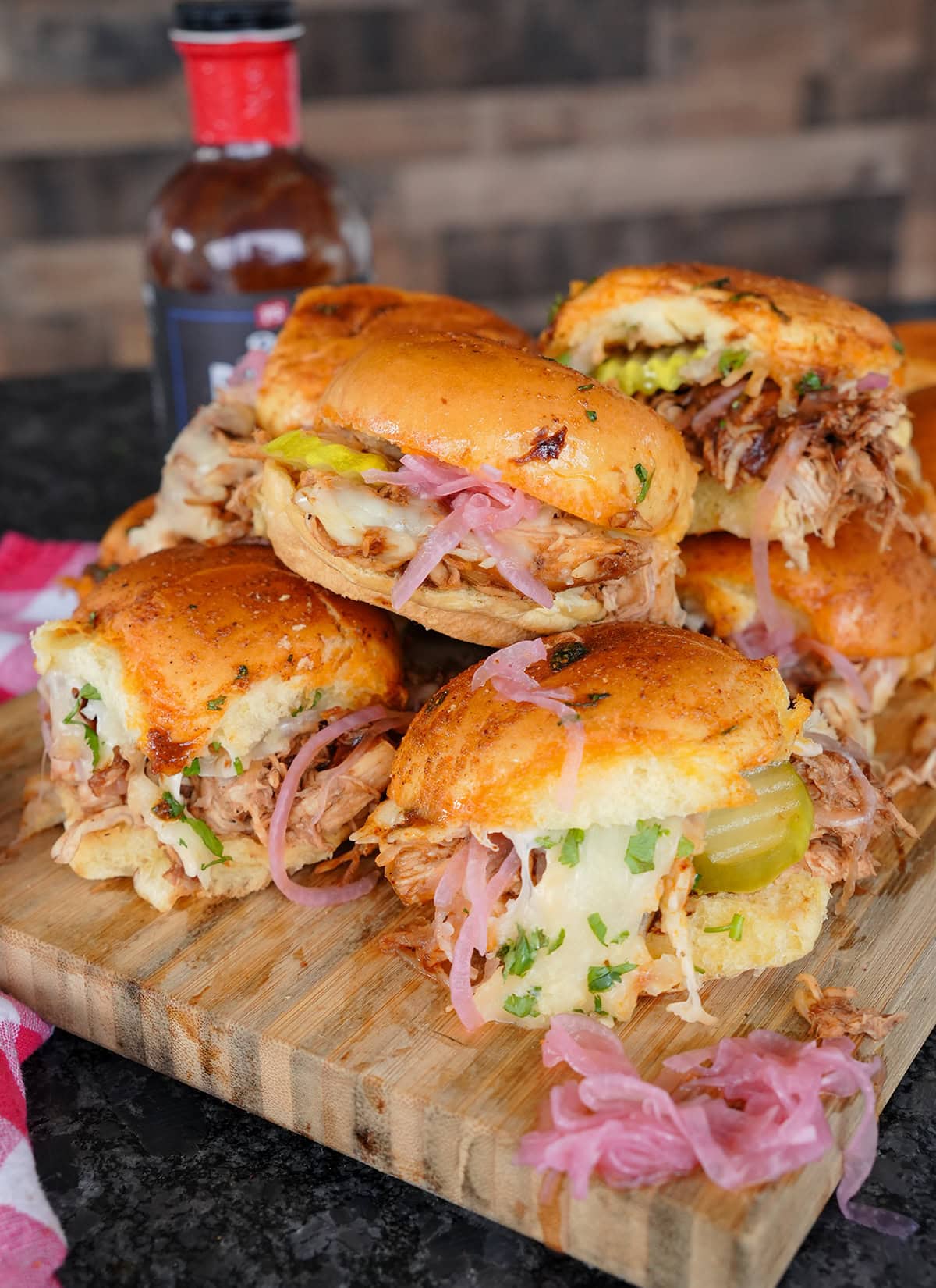 A pile of BBQ chicken sliders showing melted cheese, pickled red onions, and bbq chicken on a wooden board.