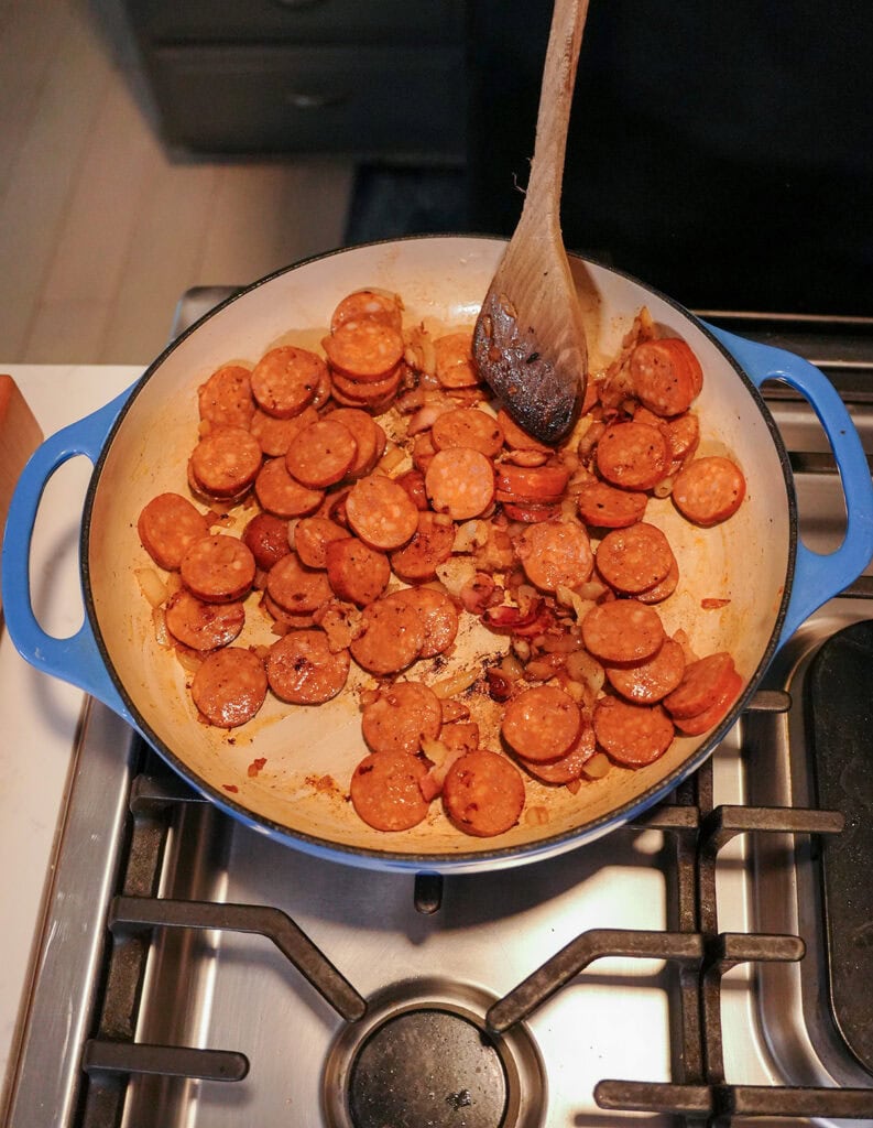 smoked sausage browning in a skillet with onion and bacon.