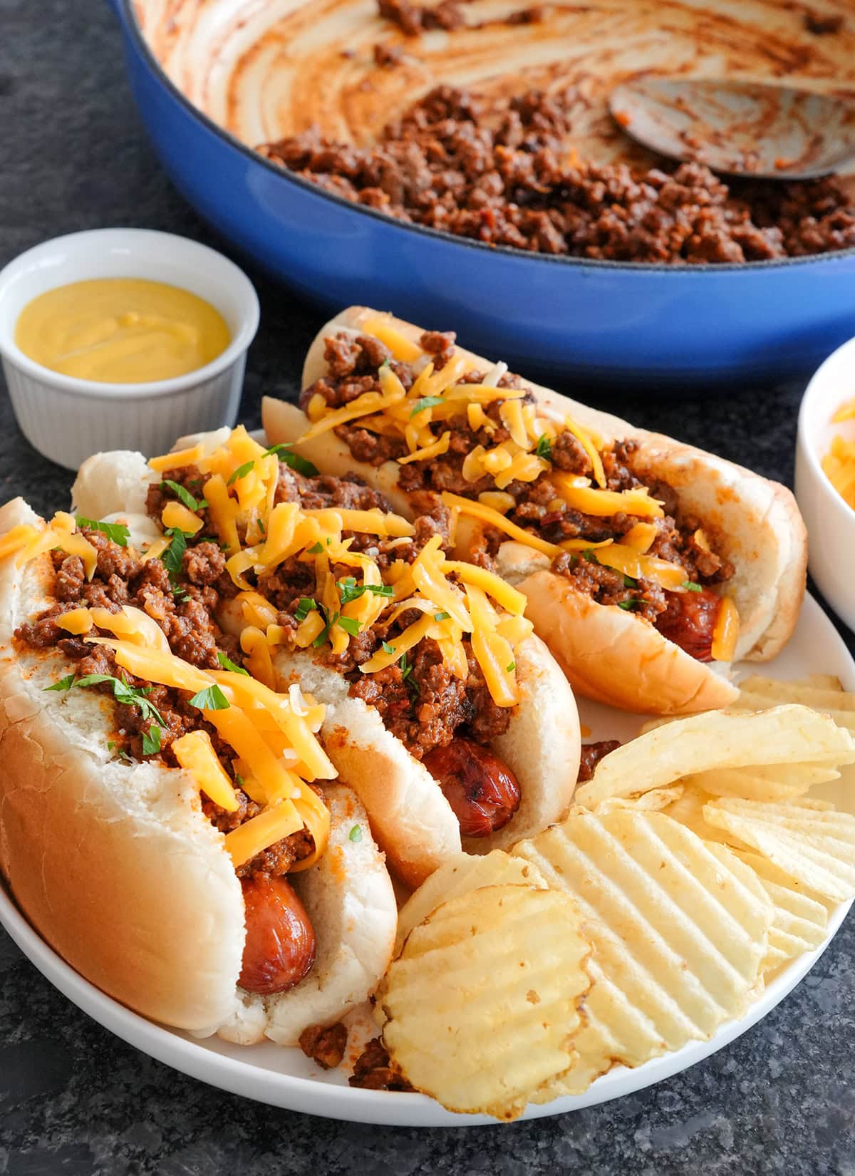 A plate of chili cheese dogs and chips with a pot of chili behind them. 