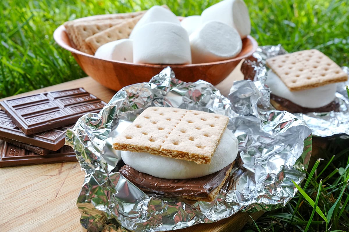 a grilled s'more in front of all the ingredients on a board set in the grass.
